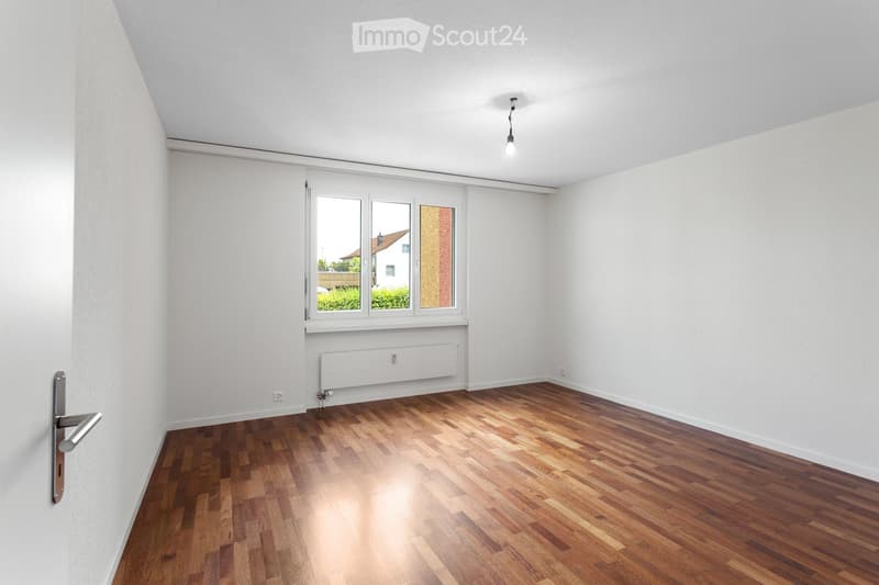 Empty room with wooden flooring, white walls, a window with blinds, a radiator, and a door handle visible.