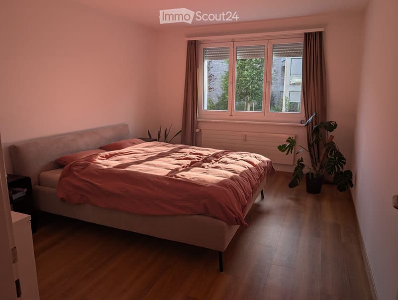 Cozy bedroom with a wooden floor, a large window with curtains, a bed with a pink duvet, a nightstand, and a potted plant near the window.