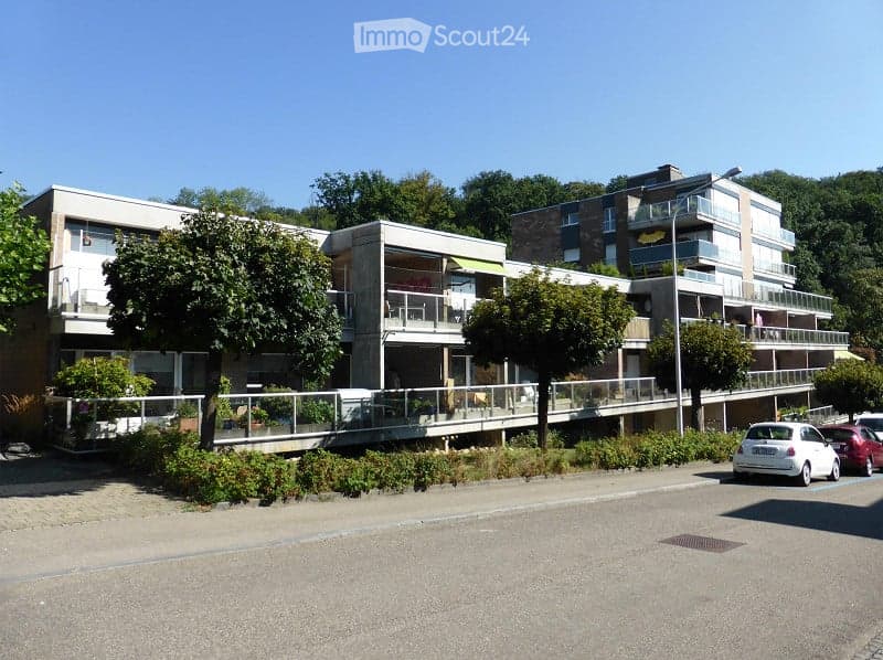 Modern multi-story residential building with balconies, surrounded by trees and parked cars.