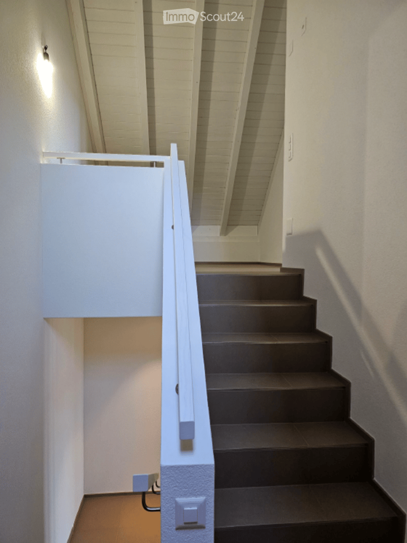 Modern staircase with dark wooden steps, white handrail, and exposed wooden ceiling beams. The walls are white, and the lighting is warm and soft.