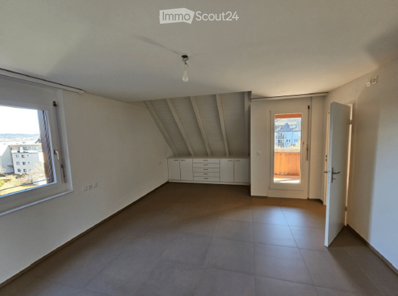 Empty attic room with exposed wooden beams, large window, built-in cabinets, and a door leading to a balcony.