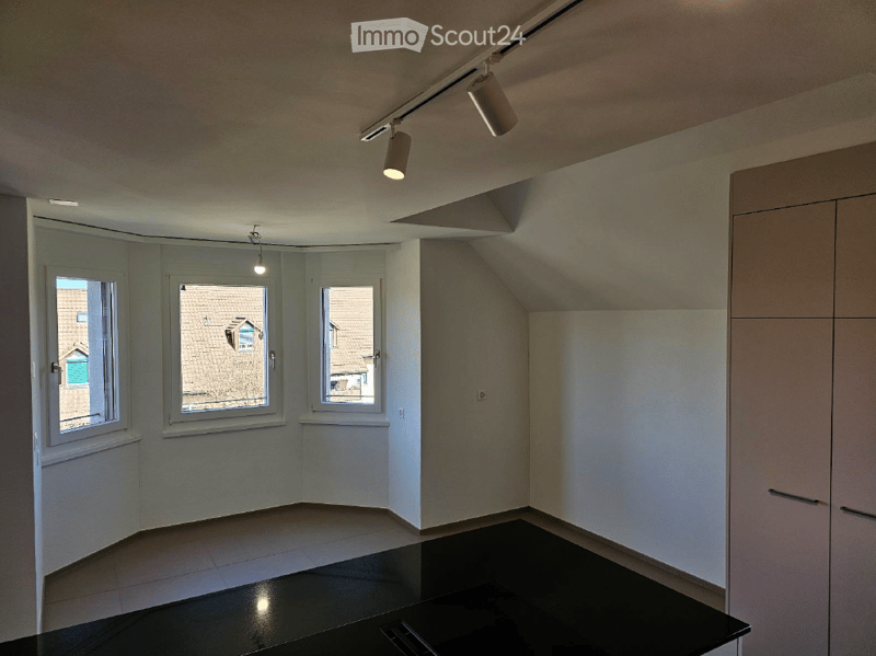 Empty room with white walls, black tiled floor, and three windows offering views of neighboring houses. Track lighting is mounted on the ceiling.