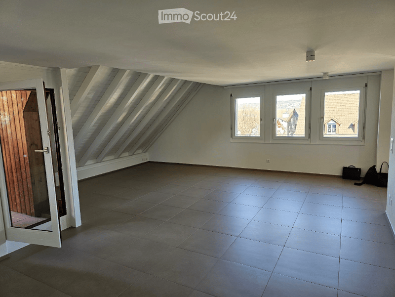 Empty attic room with slanted ceilings, large windows, tiled floor, and a wooden door leading outside.
