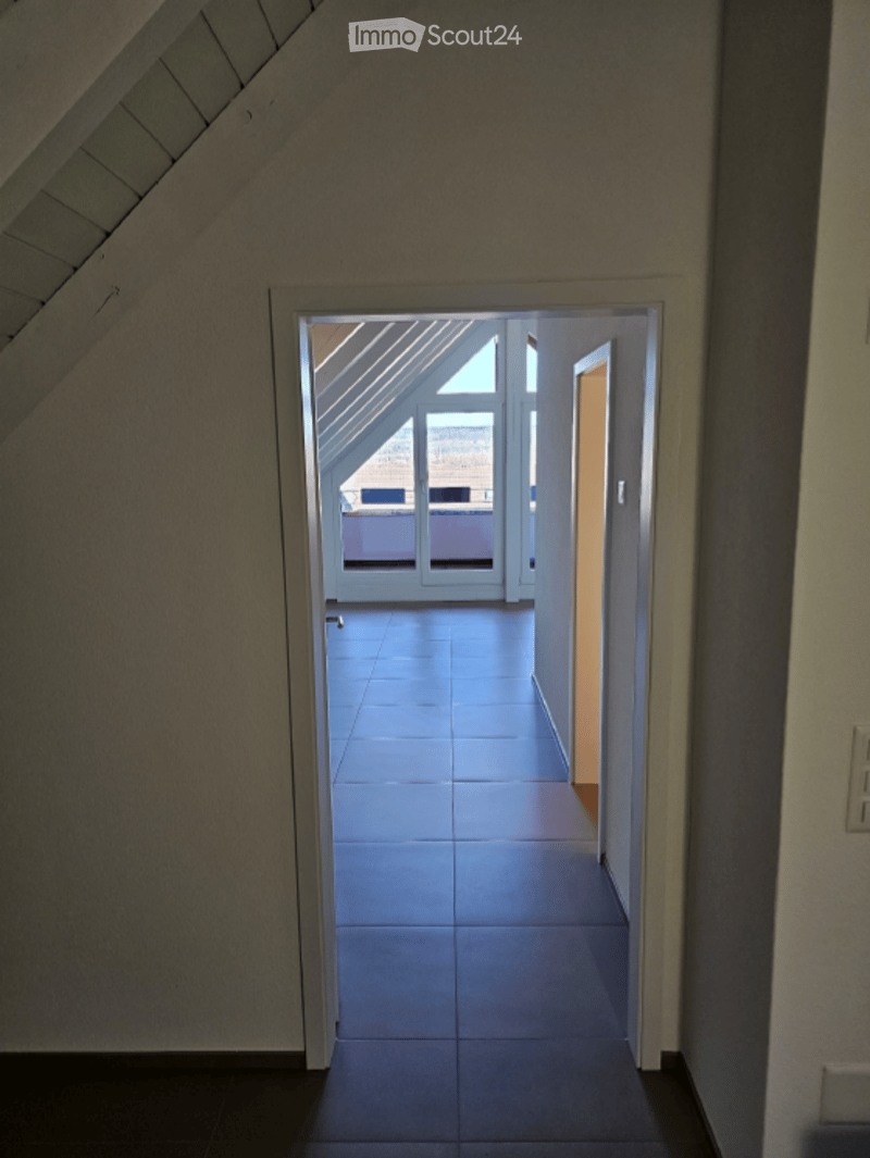 A narrow hallway with a slanted ceiling leads to a bright, open space with large windows and a tiled floor.
