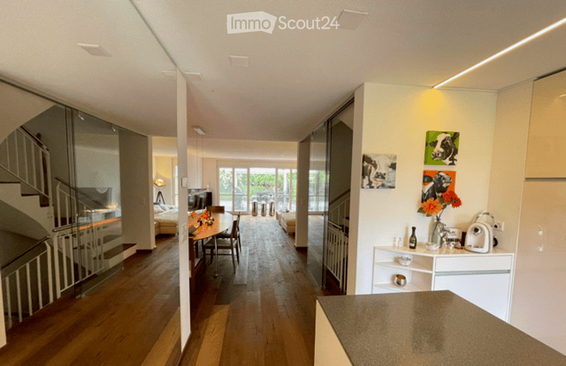 Open-plan living and dining area with wooden flooring, modern furniture including a dining table with chairs, a sofa, and a kitchen counter with a vase of flowers and framed artwork on the wall.