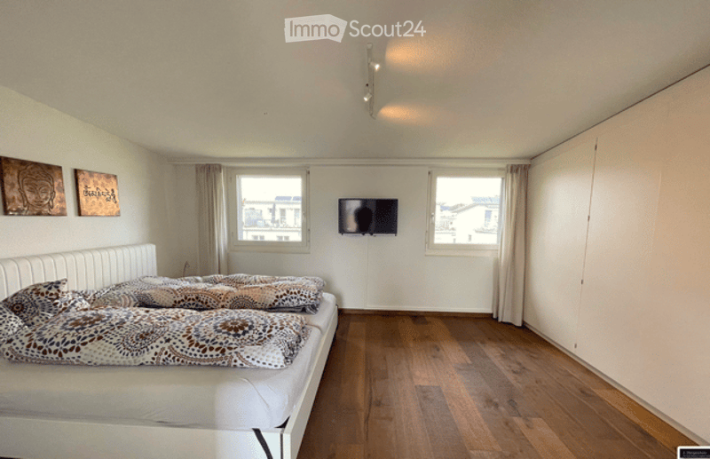 Bright bedroom with wooden flooring, large windows, white bed with patterned bedding, mounted TV, and two wall art pieces.
