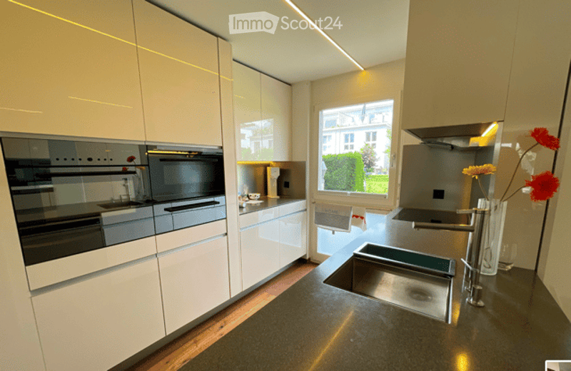 Modern kitchen with sleek white cabinets, stainless steel appliances, and a large window offering a view of greenery outside. Features include a central island with a sink and faucet, a built-in oven, microwave, and a vase with flowers on the counter.