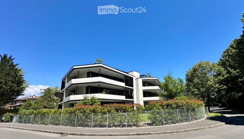 Modern multi-story residential building with balconies, surrounded by greenery and a fence.