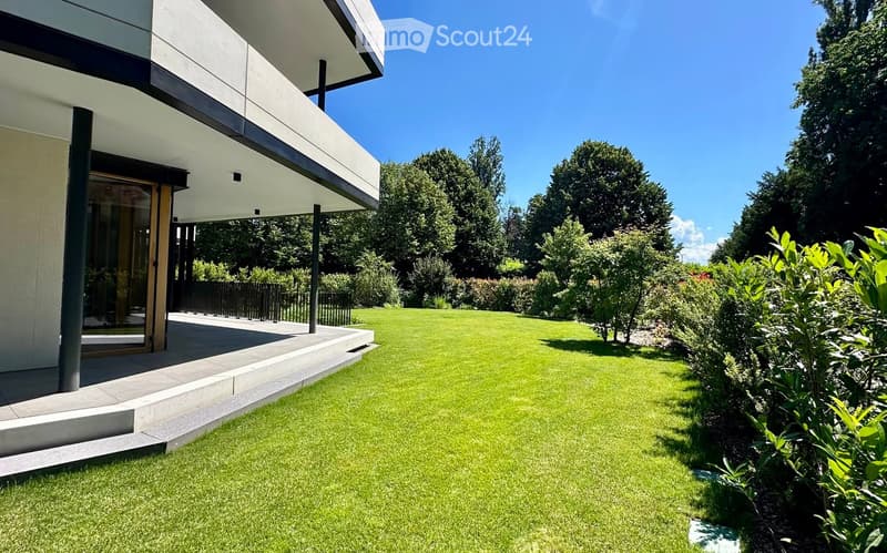 Well-maintained lawn with trimmed hedges and trees, modern house with large windows and raised ground floor visible in the background.