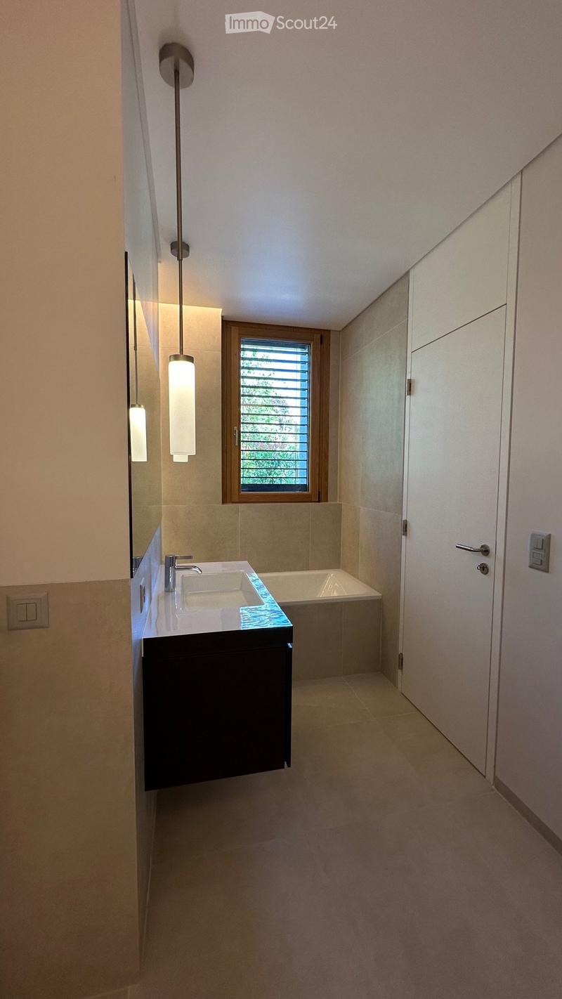 Modern bathroom with a white sink, chrome faucet, dark wooden cabinet, bathtub, and a large mirror. The room features beige tiles and a window with blinds.
