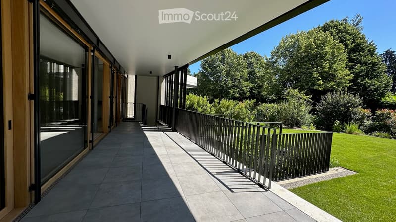 Modern balcony with large glass sliding doors, tiled floor, metal railing, and a view of a well-maintained garden with green grass and trees.