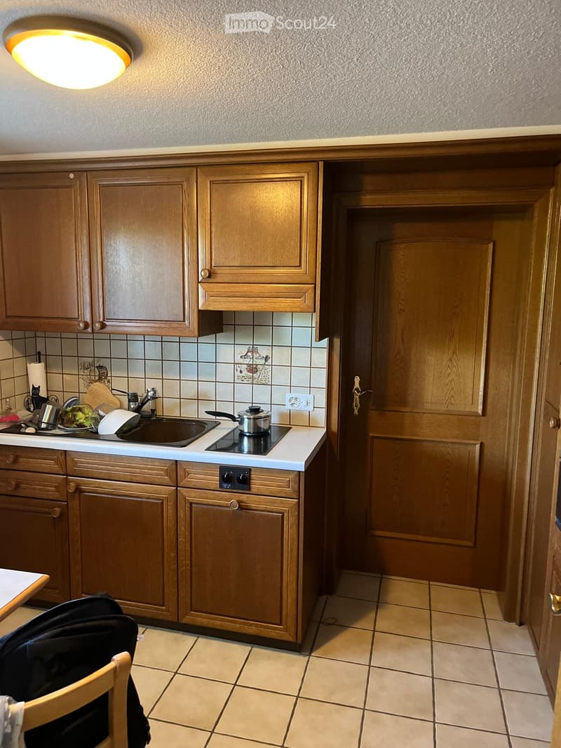 A cozy kitchen with wooden cabinets, a white countertop, and a tiled backsplash. The kitchen features a sink, stove, and various small appliances and utensils on the counter.