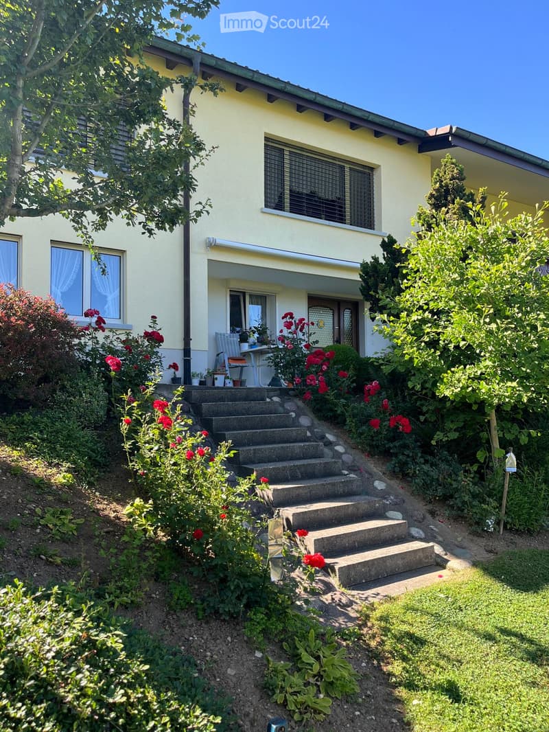 Two-story house with yellow exterior, multiple windows, and a small porch with a table and chairs. The house is surrounded by a well-maintained garden with red flowers, shrubs, and a concrete staircase leading up to the entrance.