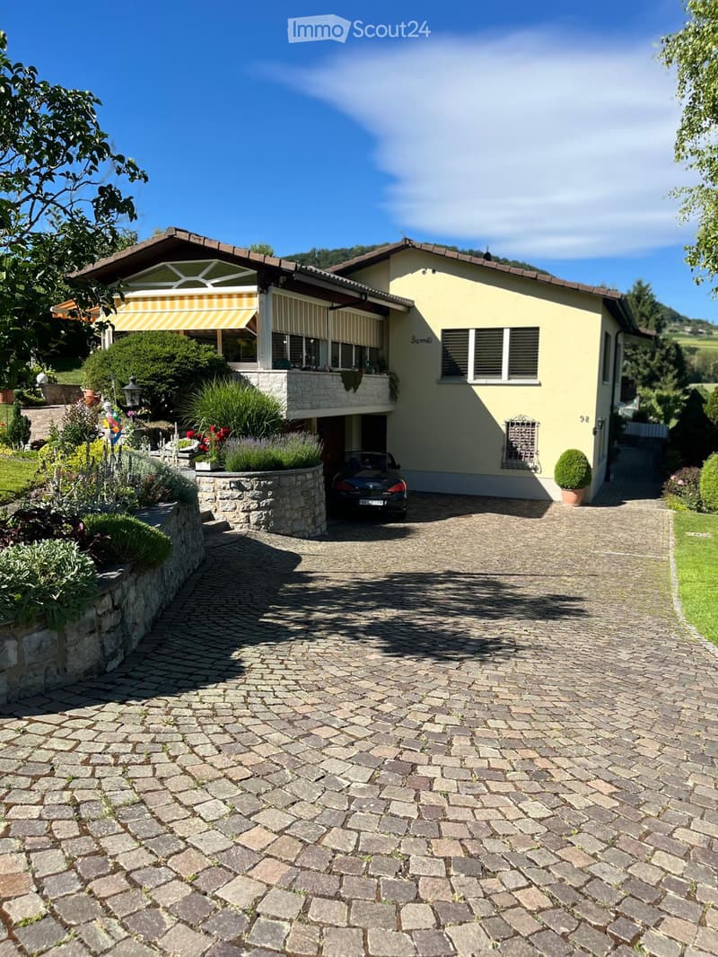 Two-story house with yellow exterior, stone driveway, garage, and garden area with plants and flowers.