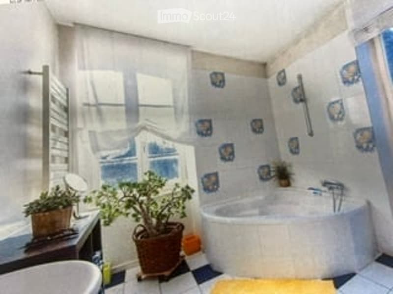 Bathroom with a white bathtub, sink, mirror, and potted plants on a small table. The walls are tiled with blue and white patterns, and there is a window with a white curtain.