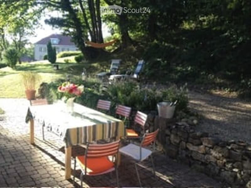 Outdoor patio area with a stone wall, wooden table with striped tablecloth, colorful chairs, potted plants, and a view of a house in the background.