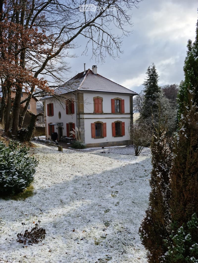 Two-story house with red shutters and a tiled roof, surrounded by snow-covered ground and trees.