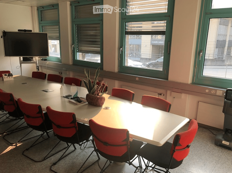 Conference room with a long table, red chairs, a potted plant, and a large screen on the wall. Windows with blinds provide natural light.