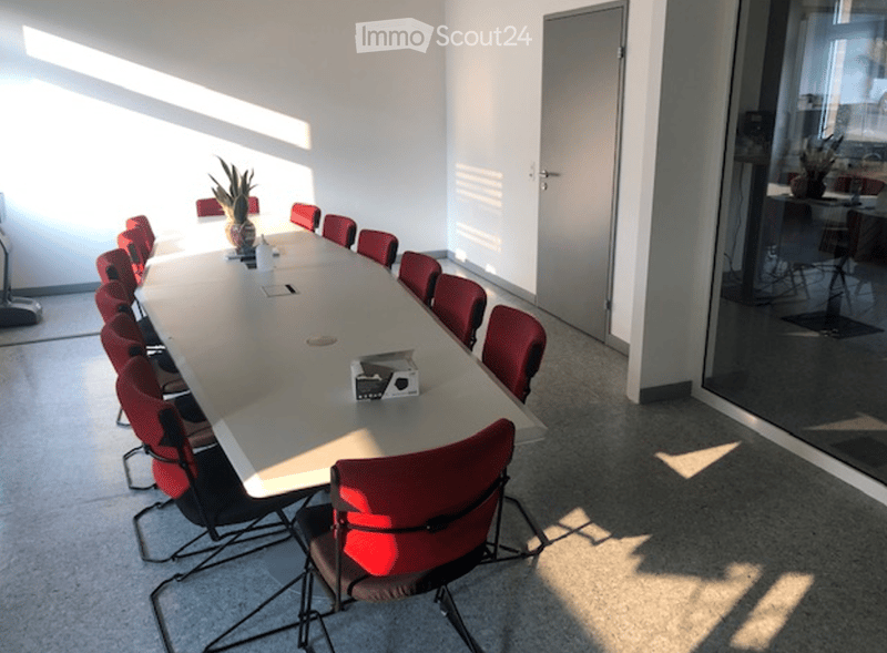 Conference room with a long table surrounded by red chairs, a vase with a plant on the table, and a glass wall offering a view of another room.