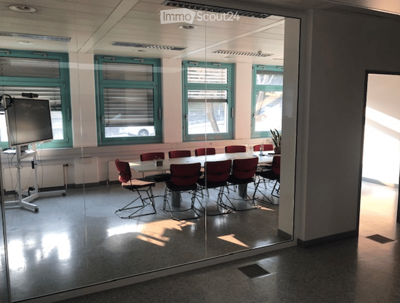 A modern meeting room with a large glass wall, a long table surrounded by red chairs, and a screen on a stand. The room has large windows with blinds and a potted plant on the windowsill.
