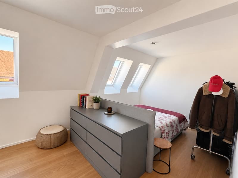 Bright attic bedroom with wooden flooring, slanted ceilings, multiple skylights, and a built-in gray nightstand with drawers. Features a bed with a patterned red and white duvet, a small round wooden stool, a woven round seat, and a coat rack with a jacke