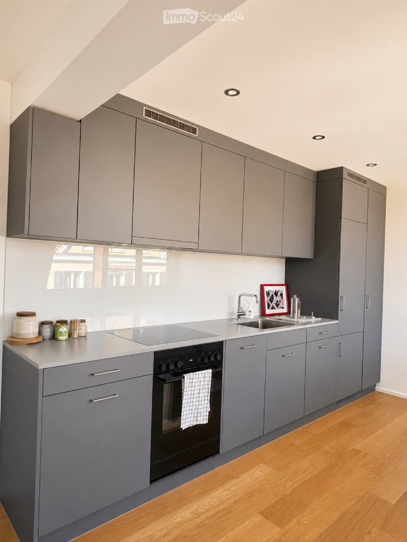 Modern kitchen with grey cabinets, a central island, an electric stovetop, and a sink. The island features jars and a framed picture, and the oven has a towel hanging from it.