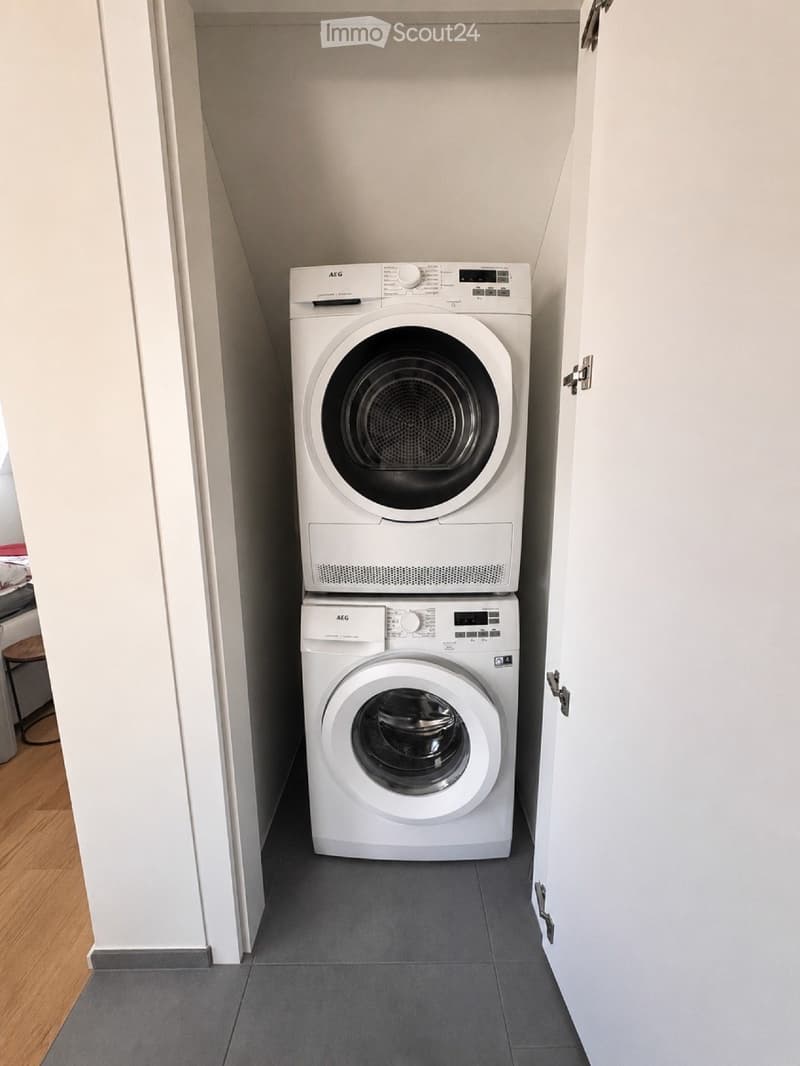Compact space with a stacked washing machine and dryer, white walls, grey tiled floor, and wooden flooring visible in the adjacent area.