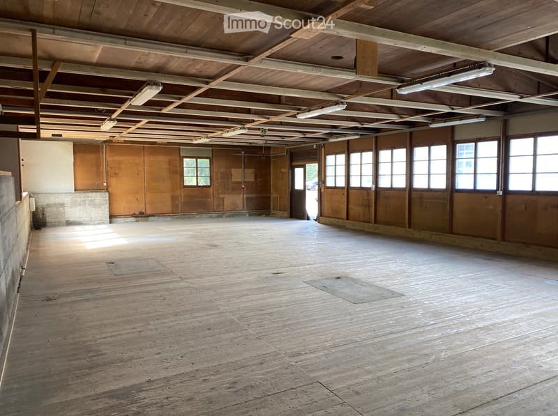 Empty room with wooden floors and exposed wooden beams on the ceiling. Large windows and doors provide natural light.