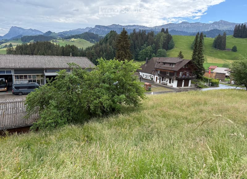 A two-story house with a garage, surrounded by lush greenery and mountains in the background. The house features a mix of modern and traditional architectural elements, with a large driveway and several parked vehicles.