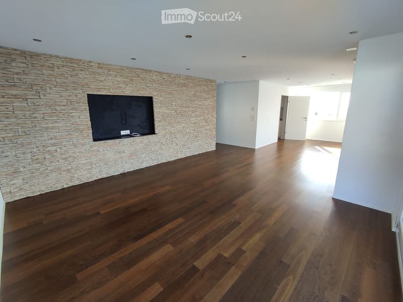 Empty room with wooden flooring and a stone accent wall featuring a mounted television. The space is well-lit with recessed ceiling lights and has white walls.