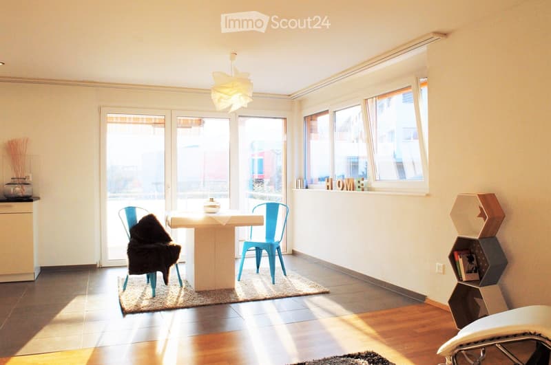Bright living space with large windows and sliding glass doors leading to a balcony. Features a white table with two blue chairs, a geometric shelf, and decorative items.