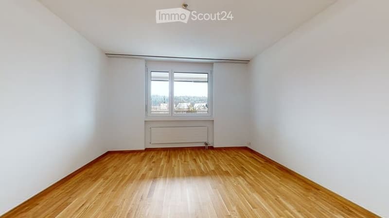 Empty room with wooden flooring, white walls, and a window with a view of trees and buildings.