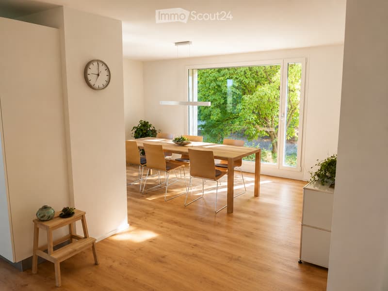 Bright dining area with wooden floor, large windows, dining table with chairs, small stool with decorative items, and a clock on the wall.
