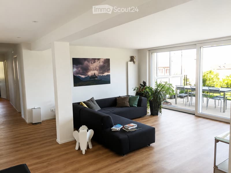 Modern living room with a dark sectional sofa, wooden floor, large picture frame, and a balcony leading to an outdoor dining area.
