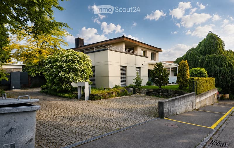Modern two-story house with a flat roof, beige facade, and a small balcony on the upper level. The house is surrounded by well-maintained greenery, including trees and shrubs, and features a paved driveway leading to a parking area.