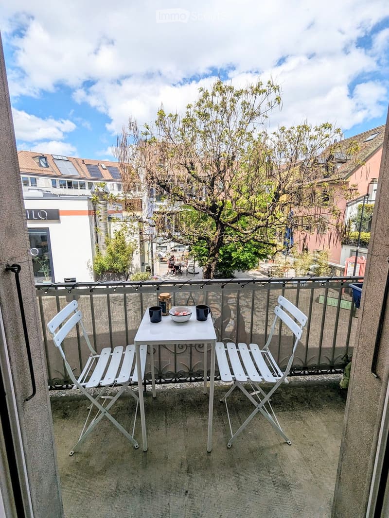 Balcony with a small white table and two white chairs, a bowl, and two cups on the table, overlooking a cityscape with trees and buildings.