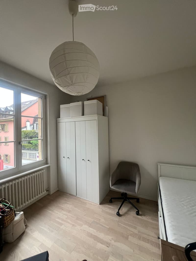 Bright bedroom with wooden flooring, white wardrobe, grey chair, and a hanging paper lantern light fixture.