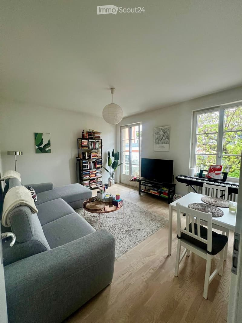 A modern living room with a grey sofa, coffee table, dining table, chairs, piano, bookshelf, and a large window with a view of greenery.