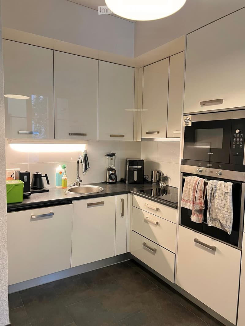 Modern kitchen with white cabinets, stainless steel appliances, and a dark countertop. Includes a sink, oven, microwave, blender, coffee maker, and various small appliances.