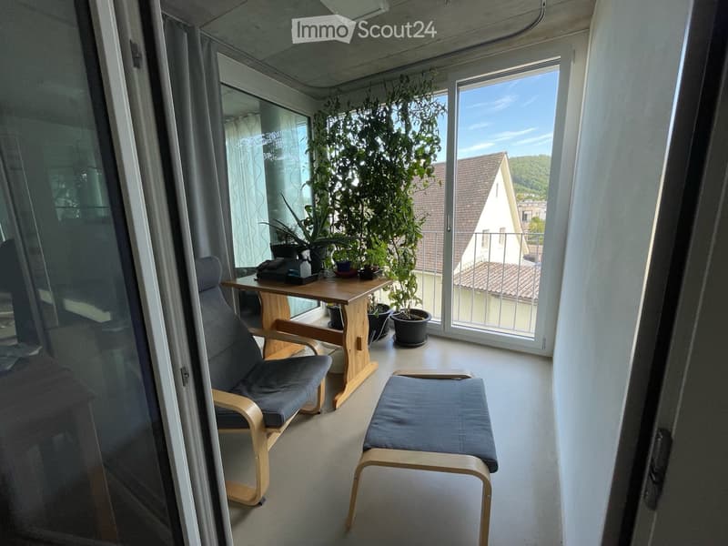 Balcony with a wooden table, two chairs, a stool, and several potted plants. Large windows offer a view of the surrounding area.