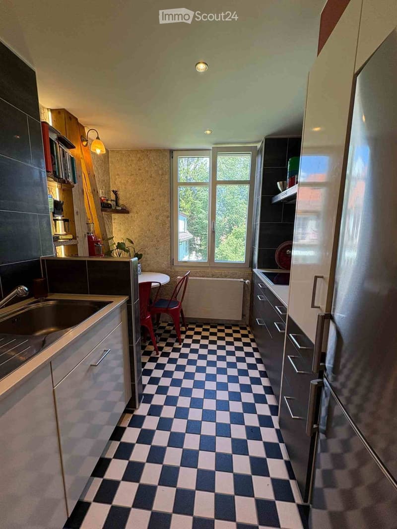 Kitchen with checkered floor, stainless steel sink, refrigerator, small round table with red chair, and built-in cabinets.