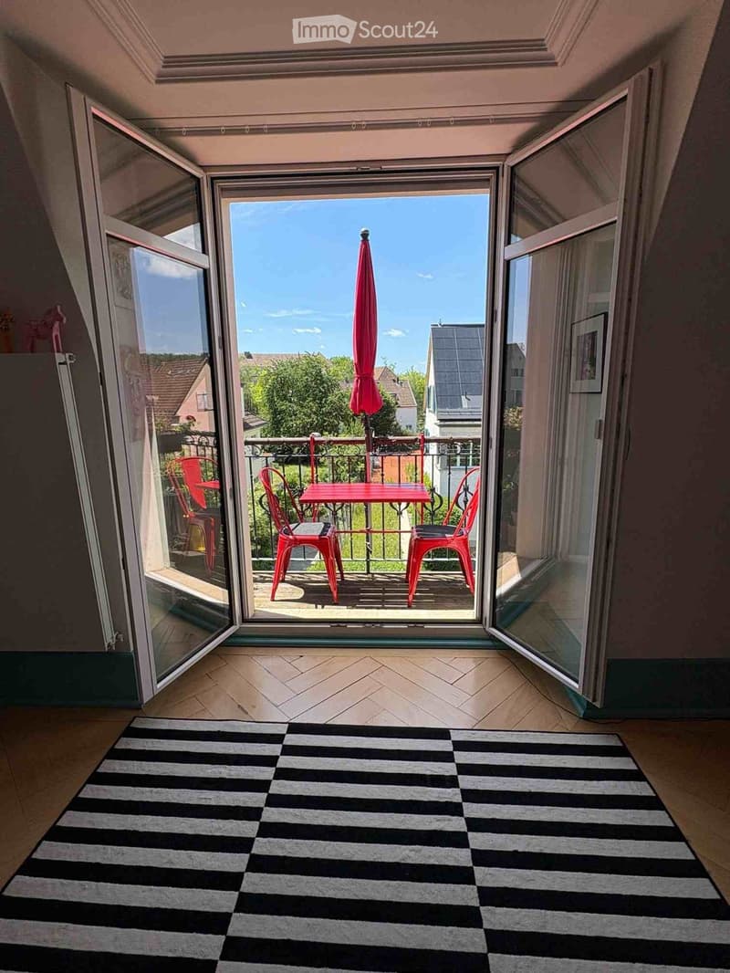 Open balcony with red table and chairs, umbrella, and a view of the neighborhood.