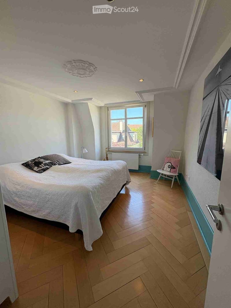 Cozy bedroom with wooden flooring, a large bed with white bedding and black and white striped pillows, a small white chair with a pink cushion, and a window with white curtains. The walls are painted in soft pastel colors, and the room features a decorati