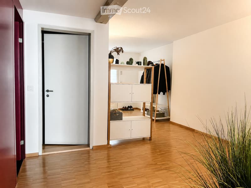 A modern hallway with light wooden flooring, white walls, and a white cabinet with shelves holding decorative items and shoes. A coat rack with jackets is visible, along with a tall plant in the foreground.