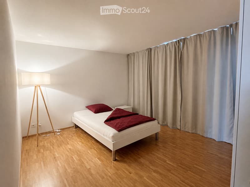 Minimalist bedroom with a white bed, red pillows, a nightstand, and a floor lamp. Light wooden flooring and neutral-colored walls.