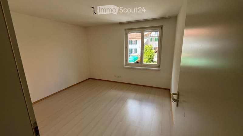 Empty room with wooden flooring, white walls, and a single window showing a view of a neighboring building and greenery.