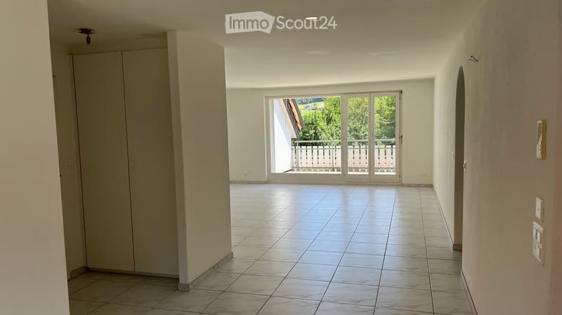 Empty hallway with tiled floor, white walls, and a large sliding glass door leading to a balcony with a view of greenery.