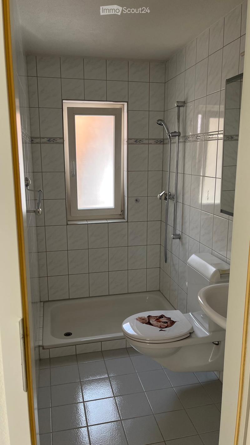 Modern bathroom with grey tiled walls and floor, featuring a bathtub, toilet with a decorative sticker, sink, and a showerhead with a handheld sprayer. A small window provides natural light.