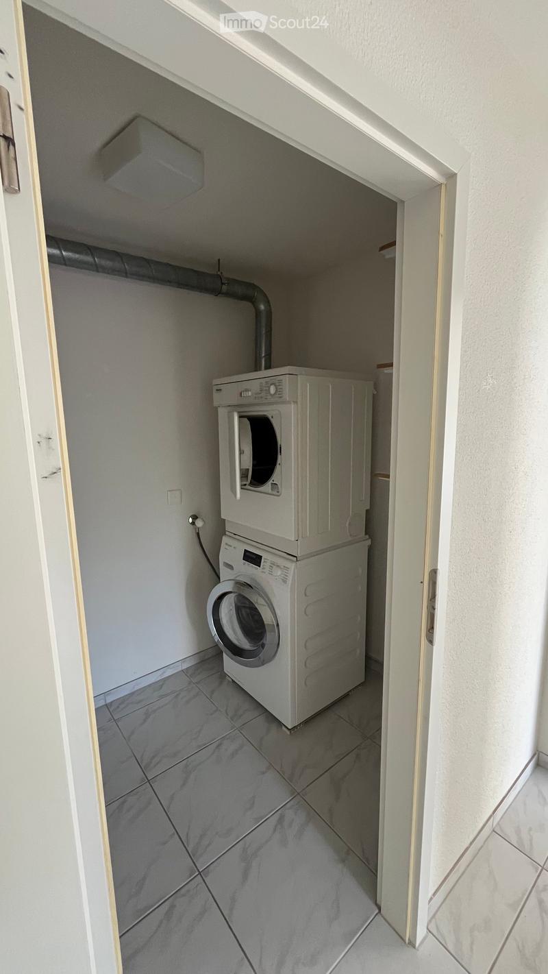 Small utility room with a washing machine and dryer stacked vertically, tiled floor, white walls, and a door leading to another area.