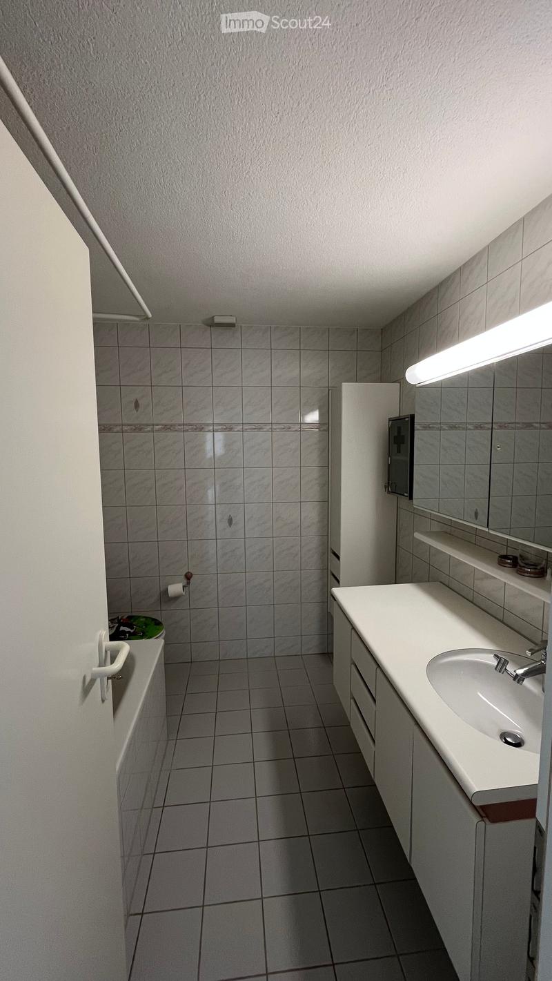 Modern bathroom with white tiled walls and floor, a white sink with a chrome faucet, a mirrored cabinet, and a toilet in the background.